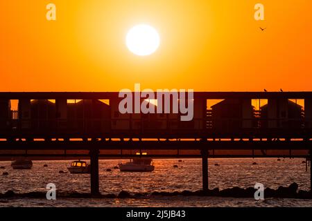 Cielo arancione chiaro con il sole che sorge sopra la silhouette del molo Herne Bar sulla costa del Kent. Dietro e sotto il molo ci sono diverse piccole barche ormeggiate nel porto. Foto Stock