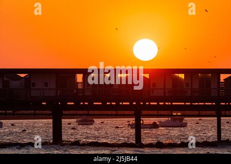 Cielo arancione chiaro con il sole che sorge sopra la silhouette del molo Herne Bar sulla costa del Kent. Dietro e sotto il molo ci sono diverse piccole barche ormeggiate nel porto. Foto Stock