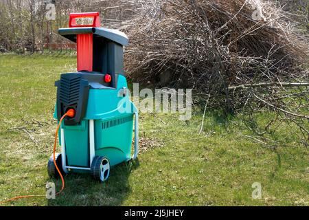 Tritadocumenti da giardino elettrico con prato stagionato e cumulo di rami di alberi tagliati sullo sfondo. Ripulisce intorno alla casa. Giardinaggio primaverile. Potatura Foto Stock