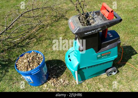 Celadna, Czechia - 04.23.2022: Dettaglio del trituratore elettrico da giardino Bosch con prato stagionato e cumulo di rami di alberi tagliati sullo sfondo Foto Stock