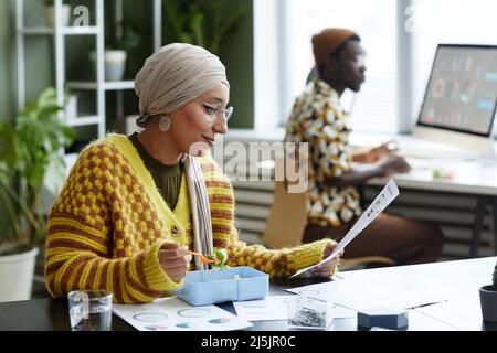 Ritratto della vista laterale della giovane donna musulmana che indossa un foulard e mangia un pranzo sano in ufficio Foto Stock