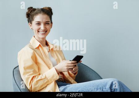 Ritratto minimale di giovane donna spensierata con smartphone, seduto contro la parete blu in ufficio e sorridente alla fotocamera, spazio copia Foto Stock