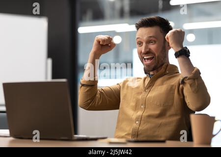 Un uomo d'affari gioioso che usa il laptop e agita i Fists seduti in ufficio Foto Stock