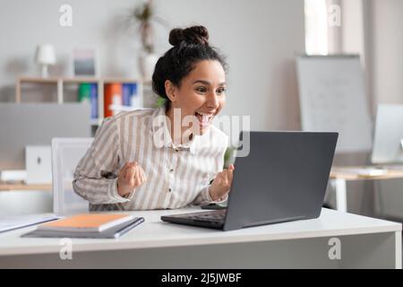 Vinci. Donna d'affari emozionale entusiasta che celebra il successo, scuotendo i pugni aggrappati e guardando il notebook in ufficio Foto Stock