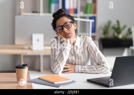 Donna d'affari stanca che dorme sul posto di lavoro mentre lavora al computer portatile in moderni interni d'ufficio Foto Stock
