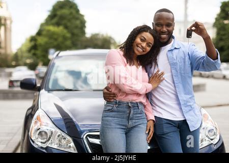 Felice coppia nera appoggiata su auto nuova mostrando chiavi auto Foto Stock