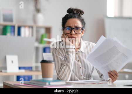 Stanca donna in ufficio che guarda la macchina fotografica e tiene documenti, seduto alla scrivania in ufficio Foto Stock