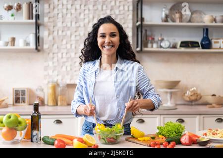 Fare pasto sano. Felice donna latina che cucinava insalata fresca, in piedi in cucina e sorridendo alla macchina fotografica Foto Stock