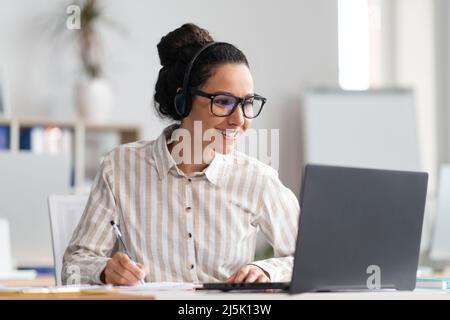 Ritratto di felice giovane donna latina indossando cuffie wireless guardando corso online, seduto al tavolo Foto Stock