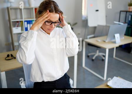 Donna d'affari stanca di mezza età che sente mal di testa da lavoro eccessivo. Ha sottolineato la lavoratrice femminile in ufficio Foto Stock