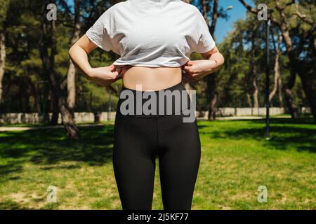 Giovane donna sportiva che indossa pantaloni yoga nero e tee bianche in piedi su una panca del parco. Foto Stock