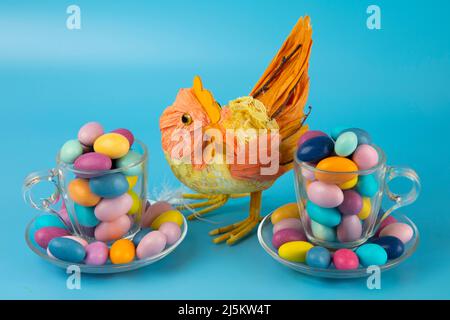 Buona Pasqua! Gallina artigianale giocattolo con due tazze trasparenti riempite di uova colorate di quaglia di Pasqua su sfondo blu Foto Stock