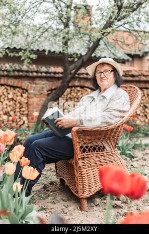 Donna anziana che si rilassa e legge un libro in giardino. Tulipani davanti a lei Foto Stock