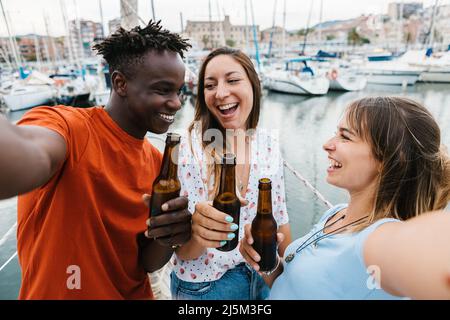 Tre amici allegri multirazziali bevono birra mentre prendono selfie in barca partito Foto Stock