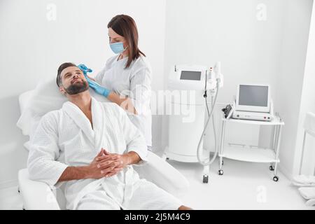 Il cliente maturo si siede in poltrona mentre il cosmetologo fa la procedura di terapia del meso nel salone di bellezza Foto Stock