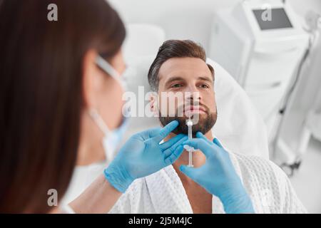 L'uomo maturo con i capelli grigi subisce la procedura di aumento del labbro con il padrone femminile in clinica Foto Stock