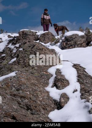 Femmina con berretto blu di beisball a piedi il suo labrador Retriever cioccolato in montagna piena di neve. Entrambi guardando l'orizzonte. Foto Stock