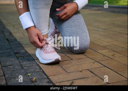 Vista ritagliata di un atleta femminile, runner che legano le scarpe sulle sue sneakers che si preparano per fare jogging lungo il tapis roulant di un parco cittadino. Sport, sollevi attivi Foto Stock