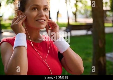Donna attiva di mezza età, sportivo indossando cuffie, maglietta rossa brillante e braccialetti bianchi in spugna, pronta per l'outdoo Foto Stock
