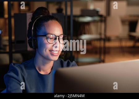 Sorridente giovane e simpatico specialista dell'helpdesk in cuffie vivavoce che risponde a domande e verifica delle informazioni sul computer Foto Stock