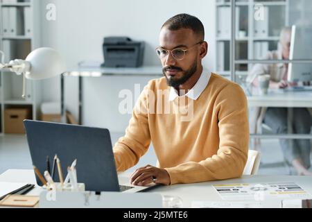 Giovane direttore Biracial concentrato con barba seduta al tavolo d'ufficio e analisi di file online su laptop Foto Stock