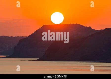 Burton Bradstock, Dorset, Regno Unito. 24th aprile 2022. Meteo Regno Unito. Il cielo si illumina di arancione al tramonto, visto da Burton Bradstock nel Dorset, mentre il sole scende dietro le scogliere di Golden Cap sulla Jurassic Coast alla fine di una calda giornata di sole. Picture Credit: Graham Hunt/Alamy Live News Foto Stock