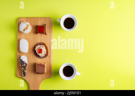 Varie torte su un asse di legno accanto a due tazze di caffè su sfondo verde. Foto Stock