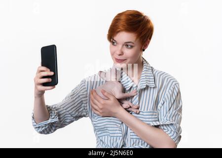 Giovane donna rossa che abbraccia il gattino domestico che dorme, usando il suo telefono cellulare e prendendo selfie alla fotocamera dello smartphone. Studio girato su sfondo bianco Foto Stock