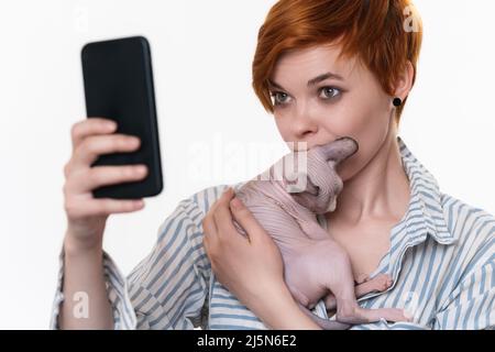 Donna che tiene e bacia il gattino, scattando foto autoritratto selfie sullo smartphone. Hipster rosso giovane adulto vestito maglia bianco-blu a righe Foto Stock