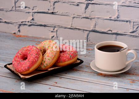 Tre ciambelle in glassa rosa e rossa su un piatto e una tazza di caffè nero su un tavolo di legno. Primo piano. Foto Stock