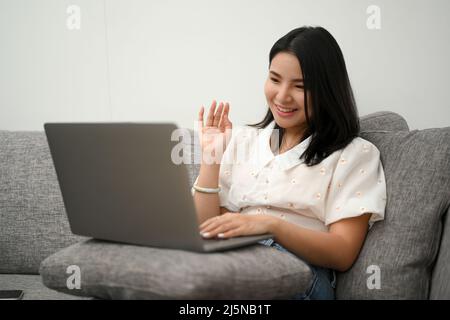 Affascinante donna millenaria asiatica si rilassa seduto sul divano, usando il computer portatile per videochiamare con la sua famiglia. Riunione online, videoconferenza Foto Stock