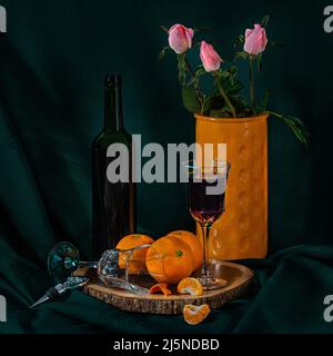 Una foto scura e suggestiva di una natura morta, una bottiglia di vino, bicchieri, arance e una bottiglia di rose. Foto Stock