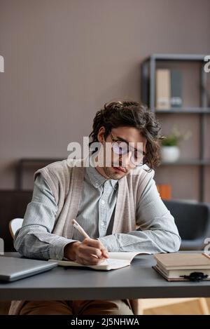 Verticale medio ritratto di bel giovane uomo adulto che indossa un elegante abito casual con occhiali seduti alla scrivania scrivere qualcosa in notebook Foto Stock