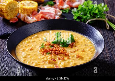 zuppa di mais con pancetta fritta croccante sbriciolata in un recipiente nero su un tavolo di legno scuro, con ingredienti sullo sfondo, primo piano Foto Stock