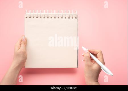 scrittura in blocco note vuoto su sfondo rosa. Mockup notepad. Mano donna con penna accanto a un blocco note vuoto vuoto. Vista dall'alto della mano che tiene la penna contro Foto Stock