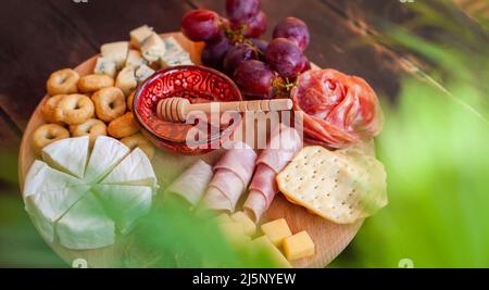 Tagliere rotondo con affettare formaggi assortiti, rosette di salsiccia, prosciutto, cracker, uva e fichi con miele. Piatto di salumi e formaggi sul tero Foto Stock