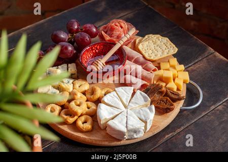 Tagliere rotondo con affettare formaggi assortiti, rosette di salsiccia, prosciutto, cracker, uva e fichi con miele. Piatto di salumi e formaggi sul tero Foto Stock