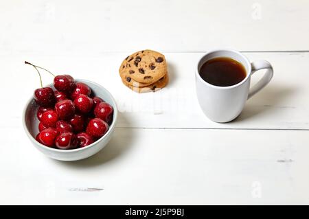 Tazza bianca con caffè, biscotti tradizionali e ciliegie dolci in una ciotola di ceramica su fondo di legno bianco, primo piano. Gustosa colazione, mattina routin Foto Stock