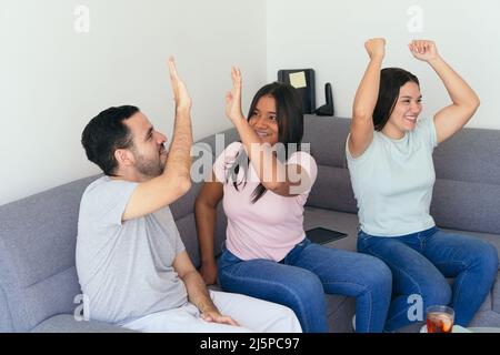 Amici che danno high-five mentre celebrano l'obiettivo di vincita. Gli appassionati di calcio potranno gustare drink e snack seduti sul divano. Stanno divertendosi a. Foto Stock