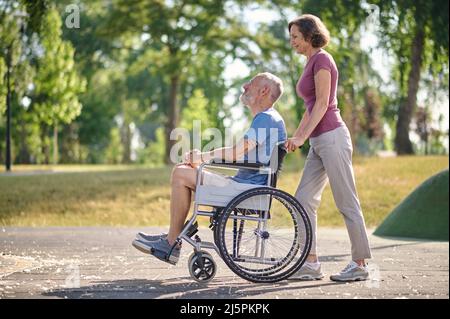 Una donna che porta il marito in una sedia a rotelle in un parco Foto Stock