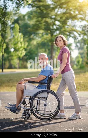 Una donna che porta il marito in una sedia a rotelle in un parco Foto Stock