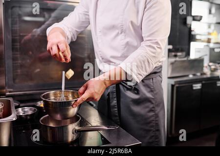 Primo piano dello chef in grembiule gettando il burro nel recipiente e fondendolo sul fornello per preparare un dessert Foto Stock
