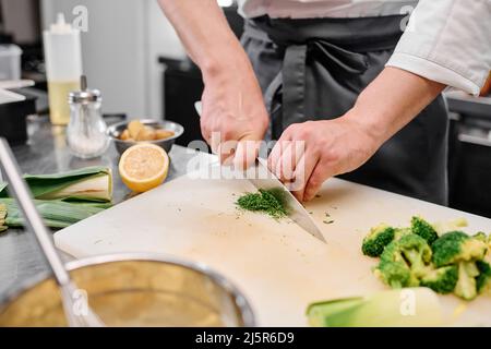 Primo piano dello chef con il coltello da cucina per tagliare i broccoli a pezzetti per il piatto sul tagliere al tavolo Foto Stock