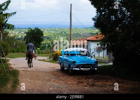 HOLGUIN, CUBA - 25 FEBBRAIO; 2020 auto classica blu e bianca parcheggiata in un villaggio sotto un albero con un cavallo e un cavaliere Foto Stock