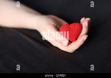 Cuore in maglia rossa nel palmo della mano femminile su un letto con biancheria da letto nera. Concetto di amore, assistenza sanitaria, maternità, donazione di sangue Foto Stock