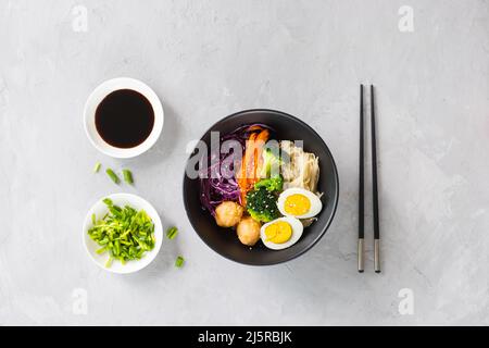 Alimentazione e dieta sane. Ciotola nera di tagliatelle asiatiche con salsa di soia, broccoli, cavolo rosso, carota, palle di gamberi, Uovo e sesamo su cemento grigio Foto Stock