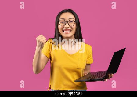 Concetto di vincita. Ritratto di signora asiatica eccitato che tiene il laptop e gesturing sì, scuotendo pugno clenched, celebrando il successo Foto Stock