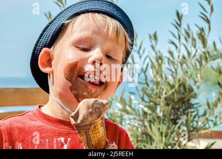 Il ragazzo biondo a panama cappello ama mangiare gelato al cioccolato che si sporca. Il bambino simpatico si siede sulla panca contro il mare in vacanza a Omis closeup Foto Stock