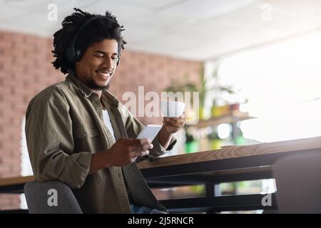 Felice giovane africano americano riccio maschio in cuffie wireless guardare lo smartphone al tavolo Foto Stock