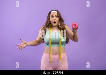 Scioccato triste giovane europeo più taglia signora in uniforme con bocca aperta e nastro di misurazione tenere ciambella Foto Stock